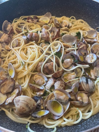 A close-up, overhead view shows a pan filled with spaghetti alle vongole. The dish features spaghetti pasta tossed with numerous whole clams, some open and some closed, mixed with chopped parsley and glistening with olive oil. The clams have varied shell patterns, ranging from mottled brown and white to darker, striated designs. The spaghetti is a pale yellow and appears to be cooked al dente. The dark grey pan has a textured surface and is filled to the brim with the appetizing pasta and seafood dish. The scene is set within Punta Ala, Italy.