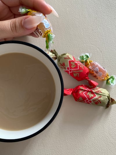 A hand with manicured, long nails holds a yellow candy wrapper with green ends and a blue and white design on it. Below the hand, a white bowl with a black rim contains a light brown liquid, likely coffee or tea. Scattered around the bowl are three individually wrapped candies, two with red and gold wrappers featuring the text "БАТОНЧИК №1" and one with an orange and yellow wrapper depicting a swirling pattern. The background is a textured, light-colored surface. The image was taken in Ostrovki, Russia.