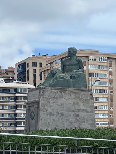 The image shows the statue of Benito Pérez Galdós, a prominent Spanish novelist, playwright, and politician, located in Las Palmas de Gran Canaria, Spain. The statue depicts Galdós seated in a contemplative pose. It is mounted on a stone pedestal, which has the name "GALDOS" inscribed on it. The buildings of Las Palmas form a background, with an apartment building visible behind the statue. Metal fencing is present on the right. The sky is partly cloudy.