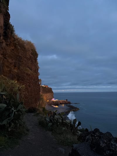 The image depicts a nighttime scene in Ponta do Sol, Portugal. The photograph is taken from an elevated position, likely a cliffside path, overlooking the Atlantic Ocean. The ocean is visible in the background, meeting a cloudy, dimly lit sky. The foreground features a rocky terrain with vegetation, including cacti and other shrubbery. A stone wall or building is situated along the cliff's edge, with faint lights emanating from the structure, perhaps indicating a settlement or buildings. The atmosphere is somber, reflecting the twilight or early evening conditions. The perspective and lighting suggest the photograph was taken after dusk.