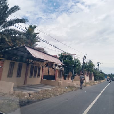 The image captures a daytime street scene in what appears to be a residential or semi-rural area of Blangmetimu, Indonesia. On the left, lush palm trees and other green vegetation provide shade for a house with a distinctive red-tiled roof, part of which forms a covered structure over a paved area. A fence constructed from light-colored concrete, accented with reddish-brown decorative pillars and intricate metalwork, separates the property from the road. A dark metal gate is also visible in the fence.

A man is depicted walking away from the camera on the dirt shoulder alongside the road. He wears a patterned short-sleeved shirt, dark pants, and a backpack. A discarded plastic bottle lies near his feet on the ground. The asphalt road features a clear white center line and recedes into the distance. In the background, several motorcycles with riders and a truck can be seen, indicating ongoing traffic. The sky is a mix of light blue and white, with scattered clouds suggesting a bright, possibly partly cloudy day. Utility lines stretch across the frame above the road and houses.