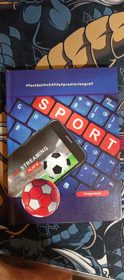The image shows a close-up of a notebook or book cover. The cover has a dark blue textured background with a graphic design of stylized blue and black shapes. Dominating the center of the cover is a graphic of a smartphone screen displaying "LIVE STREAMING" with a "PLAY" button, and two soccer balls, one white and black and the other red and white. Behind the phone and balls is a graphic of a blue keyboard with large, red, white-lettered keys spelling out "SPORT". Above the keyboard graphic, there is text that reads "#football#nfl#fifa#premierleague#". At the bottom of the cover, there is a red rectangular button with white Russian text that reads "Ежедневник", which translates to "Diary" or "Notebook". The notebook is lying flat, with its spine facing down and slightly to the right. The background outside the notebook appears to be a surface with a similar dark blue and black pattern. There is no identifiable location in the city of Kalininskaya, Russia.