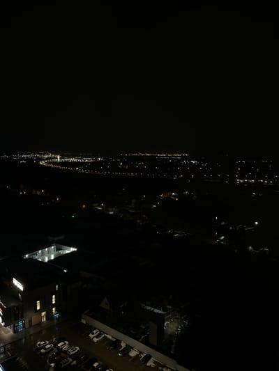 The media file presents a dark, high-angle night view of an urban or suburban landscape. The image appears to be taken from a tall building or elevated position, looking down and across a wide area.

In the distance, a long, continuous line of bright artificial lights, predominantly white and yellow, stretches across the horizon, suggesting a distant city skyline, a major road, or buildings along a river. The rest of the distant background is enveloped in darkness.

The mid-ground and foreground feature darker, more discernible structures. To the right, parts of buildings are visible, some with illuminated windows casting warm light. Below these buildings, a parking lot is evident, containing several parked cars, mostly light-colored, with some darker vehicles also present. Patches of white on the ground within the parking lot and on flat surfaces of buildings indicate the presence of snow or ice. On a dark building overlooking the parking lot, a faint, circular blue and white logo resembling the BMW emblem can be discerned.

The scene is captured at night, with artificial lights being the primary source of illumination. The presence of snow or ice strongly suggests cold weather conditions, likely winter. No human activity or movement is visible, contributing to a serene and still atmosphere. Other than the recognizable BMW logo, no distinct or readable text is visible anywhere in the image.
