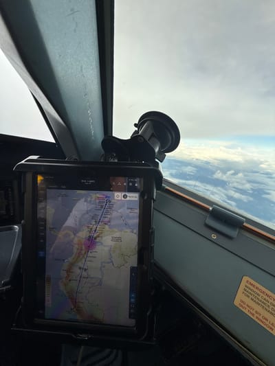 Here's a description of the image:
The image is taken from the cockpit of an aircraft, likely during flight, as evidenced by the view of the sky and clouds through the windshield. A tablet is mounted on the dashboard using a suction cup mount. The tablet displays a flight navigation application, showing a map of the Caribbean region, specifically featuring flight routes and waypoints. Text on the screen indicates a route from SPJC (Jorge Chávez International Airport in Lima, Peru) to MDPC (Punta Cana International Airport, Dominican Republic). Place names visible on the tablet include Jamaica, Colombia, Ecuador, Venezuela, and Curaçao. Additional information on the screen includes flight planning details, waypoints, and navigational aids, indicating a "High IFR" flight plan. A yellow warning label is seen in the upper right, cautioning against leaning out the window.