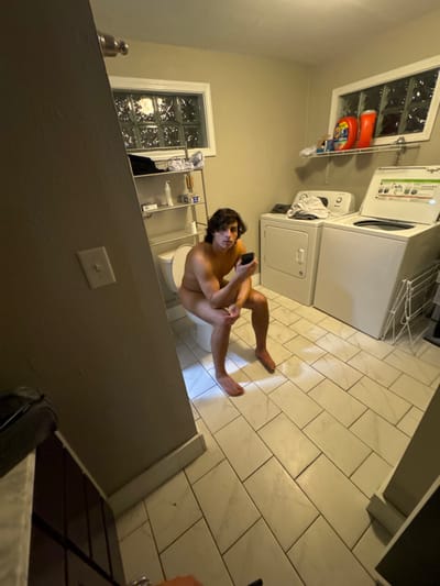 The image displays an indoor scene within a combined bathroom and laundry room. A nude male, exhibiting a fair complexion and dark, wavy, medium-length hair, is seated on a white toilet in the left-midground. He holds a dark-colored smartphone in both hands, positioned in his lap, and looks directly at the camera.
The room's walls are painted a light beige. The floor is covered with rectangular white tiles arranged in an offset pattern, separated by dark grout lines. Two horizontal rectangular windows, each fitted with opaque glass block panes, are situated on the back wall above the fixtures.
To the left, directly above the toilet, is a multi-tiered chrome-colored shelving unit containing various toiletries, including bottles and a roll of toilet paper. To the right of the toilet, against the back wall, are a white front-loading clothes dryer and a white top-loading washing machine with its lid open. Above these laundry appliances, a white wall-mounted shelf holds several large plastic bottles, some appearing to be laundry detergents. A white, collapsible drying rack is partially visible on the far right. Overhead artificial lighting illuminates the scene.
No specific external identifiers are present within the image to determine a precise location within the city of South Bend, United States; the scene depicts a residential interior.