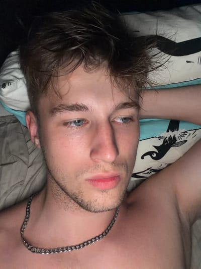 A close-up, high-angle selfie shows a fair-skinned young man with light brown, tousled hair lying on his side. He has blue eyes, fair eyebrows, a lightly stubbled jawline, and pink lips. He is wearing a silver chain necklace. He is lying on a pillow with a black and white graphic of a bird on a light blue background, and a beige comforter or blanket. The background is dark. The image is undated and unlocated, and no information about the city of Vanino, Russia is available.