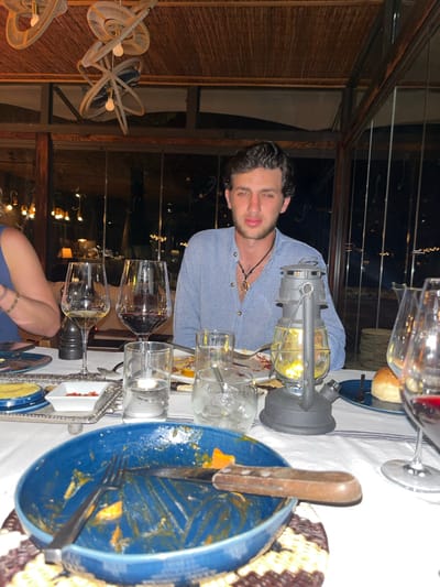 A man sits at a dining table in what appears to be an outdoor or semi-outdoor restaurant, likely in Livingstone, Zambia, given the described setting. The restaurant has a ceiling constructed of dark wooden elements and fans. The man is wearing a light blue shirt and is looking towards the viewer. On the table, a vintage-style lantern is visible, alongside wine glasses, drinking glasses, plates of food with remnants of a meal (one of the plates is blue and contains remnants of orange-colored food), cutlery, and small serving dishes. A piece of bread or roll can be seen. The table is covered with a white cloth, potentially with stripes. Through an opening in the structure, there are distant lights and a cityscape. A woman's arm is also visible.