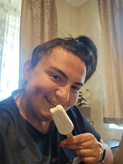 This image captures an individual indoors in Rudnik, Bulgaria, engaged in consuming a white popsicle or ice cream bar. The subject, a person with dark hair styled in a bun, features a visible dimple on the left cheek and is smiling broadly, revealing teeth. They are wearing a dark, short-sleeved t-shirt, a silver-toned chain necklace, and a metallic wristwatch on their right wrist. Pink nail polish is visible on the fingers of the right hand, which holds the wooden stick of the ice cream. The background on the left reveals a window covered by a sheer white lace curtain with a floral pattern, allowing bright natural light to enter. To the right, a drawn light brown or beige curtain with a distinct pattern is visible. A potted plant with dark green leaves is situated behind the individual's right shoulder. The wall behind the subject appears to be a light, neutral color.