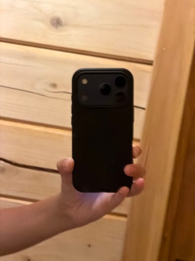 The image shows a close-up of a person's hand holding a black smartphone horizontally. The phone is positioned in front of a wooden wall, which appears to be made of vertical planks. The lighting suggests it is daytime, with natural light illuminating the scene.
The smartphone has a prominent camera module on the back, featuring three lenses arranged in a triangular pattern and a circular sensor in the middle. The hand holding the phone is positioned behind it, with fingers wrapped around the edges, suggesting the person is either taking a photo or recording a video. The skin tone of the hand indicates it belongs to a person with lighter skin.
The background consists of the wooden wall, which has a natural, slightly rough texture with visible wood grain and knots. The planks are joined vertically, creating a rustic or cabin-like aesthetic. Above the wall, there's a glimpse of a wooden ceiling or beam, also made of natural wood, suggesting an indoor setting that might be a home, cabin, or similar structure.
There are no visible text elements in the image. The activity taking place is the act of holding and using a smartphone. The overall impression is a casual, everyday moment captured indoors, with a focus on the object being held and the textured wooden background. The setting, with its wooden elements, could be interpreted as a cozy or natural environment. The image does not convey any specific emotions beyond the neutral act of using a phone.