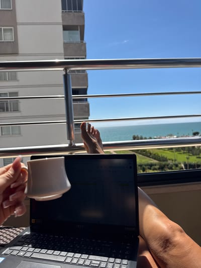 A person is seated on a balcony with a laptop and a mug of coffee, overlooking a beach and an urban landscape, presumably in Erdemli, Turkey. The view through the metal railing shows a coastline with light-colored sand, turquoise water, and a green space adjacent to the shore. The laptop displays a spreadsheet or grid-like document, and the person's leg is visible on the right side of the frame. The foreground includes the person's hand holding the mug and a glimpse of apartment buildings across from the balcony.