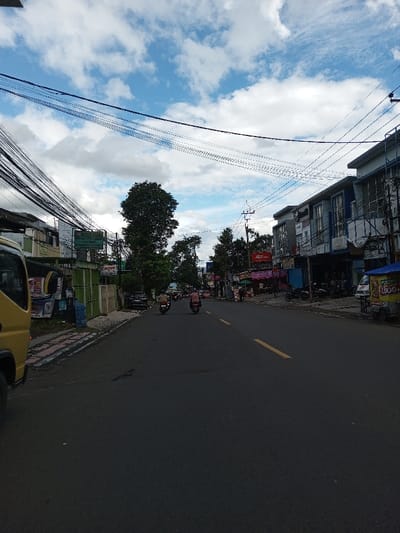 This image depicts a street scene under a partly cloudy sky, viewed from a slightly elevated position looking down a paved asphalt road in the city of Serpang Shalli, Bhutan. The dark gray roadway features two broken yellow lines marking its center. In the mid-ground, two individuals are seen operating motorcycles, traveling away from the camera; additional vehicles are visible further down the street.
On the left side of the frame, a yellow truck's front section is visible in the foreground, parked alongside a patterned sidewalk with red and gray pavers. Behind the sidewalk, a green barrier or fence runs parallel to the road, with various posters and advertisements affixed. Further back, buildings and dense green trees line the street, interspersed with numerous overhead power and communication lines that appear heavily bundled. A sign with the word "Canon" is discernable amidst the foliage.
The right side of the road features a sidewalk fronting multiple multi-story commercial buildings. A prominent red sign for "J&T Express" is mounted on one building. Other businesses display various signs, mostly unreadable. Several motorcycles are parked on the sidewalk in front of these establishments. A light blue vendor cart with a yellow canopy, displaying "1.000," is positioned on the sidewalk in the lower-right foreground. Indistinct figures are visible on the sidewalks near the businesses. The sky above is characterized by scattered white cumulus clouds against a blue background, crisscrossed by overhead utility cables.