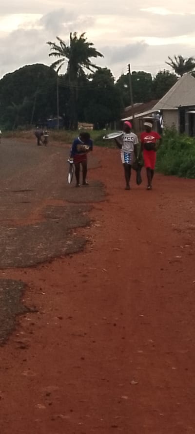 This image depicts an outdoor scene on a dirt road in Adikpo, Nigeria, under an overcast sky. Three individuals are walking on the road, with the foreground dominated by reddish-brown dirt and patches of weathered asphalt. To the left, a person in a blue and yellow long-sleeved shirt and a red skirt is walking and holding a bicycle wheel. In the middle, a person wearing a white t-shirt with "FCC" printed on it and a patterned skirt is carrying a round metal tray. To the right, a person in a red t-shirt and a red skirt is walking with their hands behind their back, possibly carrying something. In the background, trees, including palm trees, line the side of the road, with some buildings and power lines visible. The overall impression is a rural or semi-rural setting.