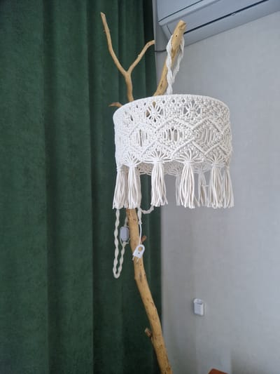 A macrame lampshade hangs from a natural branch that forms the structure of a floor lamp. The lampshade is white and features intricate knotwork with tassels hanging from its bottom edge. A white cord with an inline switch and plug dangles from the branch, connected to the lamp. The lamp stands next to a dark green curtain. In the background, a portion of a white air conditioning unit is visible mounted on a light gray wall. A small white holder is affixed to the wall. The scene appears to be an indoor domestic setting in the city of Podsteрki, Russia.