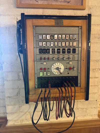 The image shows a vintage switchboard telephone mounted on a white brick wall, possibly in Sława, Poland. The switchboard is made of wood and metal, with multiple rows of buttons, knobs, and a rotary dial. A black telephone handset is attached to the side of the unit. Several cables are connected to the bottom of the switchboard, hanging down towards a wooden surface below. The environment appears to be indoors, with a framed picture visible on the wall above the switchboard. The lighting suggests it is daytime, with natural light illuminating the scene.