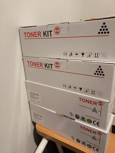 A vertical, medium shot captures a stack of four white cardboard boxes containing toner cartridges. The boxes are arranged one on top of the other, leaning slightly to the right. The top three boxes prominently display the words "TONER KIT" in red capital letters. The top two boxes also feature a red circular logo with "NEU" inside, and a graphic of nine black dots arranged in a triangle. Below the "TONER KIT" text on these boxes, there are various shipping and safety icons, including "CE" marking, symbols for fragility, rain, temperature ranges, and upright handling. The third box down has "TONER KIT" in red and the "NEU" logo. The fourth box, partially visible at the bottom, also has "TONER KIT" in red and the "NEU" logo, along with "NON OEM PRODUCTS" and "CE" marking. The third box from the top has additional text detailing "Premium Toner Cartridge" and "CPHT305M". The background consists of a textured white wall on the left and a wooden surface at the bottom, suggesting an indoor storage or retail environment. The lighting is ambient, casting soft shadows. The image is taken from a slightly upward angle.