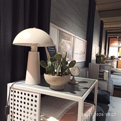The image depicts an interior scene, likely a showroom or retail space, featuring furniture and decorative items. In the foreground, a white mushroom-shaped lamp stands on a white side table with a perforated front panel. The table surface is a dark gray, wood-grain pattern. On the table, next to the lamp, is a light-colored ceramic pot containing a cactus plant. A small, round analog clock with a gold face and hands is also present on the table. Behind the table, a dark curtain hangs, and further back, a wall is clad in dark gray wood planks. Several framed posters are visible on the wall, one of which features text in Italian that translates to "Looking for the perfect mattress?". In the background, rows of mattresses are displayed, and some shelving units with perforated panels are present. The overall ambiance suggests a modern and organized retail environment. The timestamp "24.07.2026 10:07" is superimposed on the lower right portion of the image, indicating the date and time the photograph was taken. There are no people visible in the image. The scene is well-lit, suggesting overhead artificial lighting. This scene is located in Modugno, Italy.
