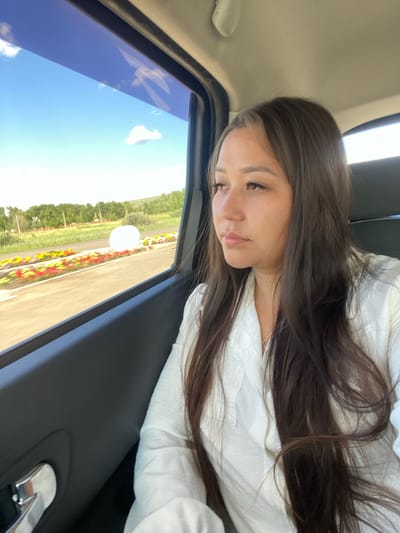 A young woman is seated in the passenger seat of a car, looking out the window. The woman has long, dark brown hair and is wearing a white button-up shirt. The car's interior is visible, with the dark gray door panel and a silver door handle in the foreground. Outside the window, a bright blue sky with scattered white clouds is visible, along with a lush green landscape of trees and a field of colorful flowers lining a road. The setting appears to be a scenic area, possibly in or near Isyangulovo, Russia, given the rural scenery. The image captures a moment of quiet observation as the woman travels.