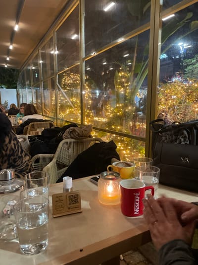 This media file captures a cozy evening scene within what appears to be a glass-enclosed cafe or restaurant in Chaidari, Greece. The time of day is night, indicated by the darkness outside and the numerous warm white string lights adorning trees and bushes, which create a festive and inviting ambiance.
In the immediate foreground, a light-colored wooden table is set with various items. To the right, a person's hands, wearing a ring, are clasped together. Central to the table are a red mug prominently displaying the "NESCAFÉ" logo, a yellow mug, and several clear drinking glasses, some appearing to hold water. A lit candle in a glass jar provides a soft, warm glow. A black handbag, possibly bearing initials such as "LV," rests on a chair next to the table. Further to the left on the table, a wooden stand with a QR code is visible, featuring the text "SCAN REVIEW" and "LOOK," likely for customer feedback or menu access.
Several people are present in the setting. One individual's head and upper body are partially visible in the background, seated at a table. Another person is partially seen in the lower left, wearing a leopard-print garment. The chairs have distinctive light-colored frames, possibly wicker or metal. Through the large glass panels forming the cafe's enclosure, the abundant string lights outside are a focal point, creating reflections on the glass that add to the visual depth. The overall atmosphere is relaxed and social, suggesting patrons are enjoying drinks and conversation.