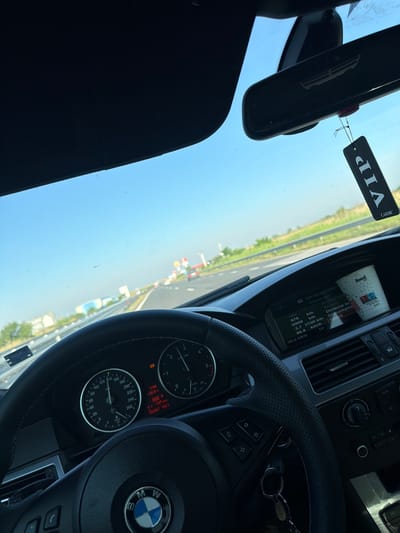 The image is taken from the driver's perspective inside a car, looking forward through the windshield. The car is on a highway, indicated by the road markings and barriers. The sky is clear and blue, suggesting a sunny day. In the foreground, the steering wheel, dashboard, and instrument cluster are visible. The instrument cluster shows a speedometer reading of approximately 90 km/h and other engine diagnostics. A "VIP" air freshener hangs from the rearview mirror. In the background, other vehicles can be seen on the highway, and the landscape appears to be rural with fields and some industrial buildings visible in the distance. The presence of a road, highway infrastructure, and the general appearance of the surroundings are consistent with a location on the outskirts of a city or town. Considering the prompt specified the city of Benkovski, Bulgaria, this scene depicts a car traveling on a highway near or leading to Benkovski. There are no people clearly visible within the frame.