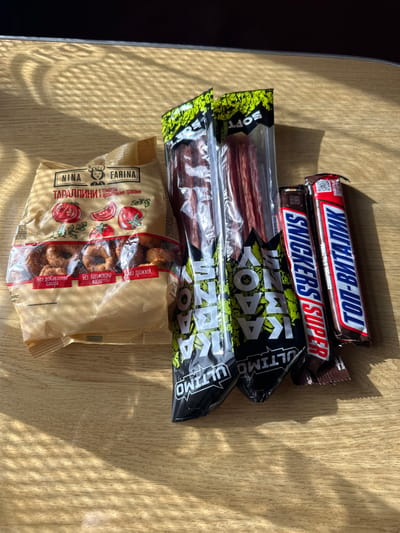 The image shows a selection of food items on a wooden surface, possibly a table. There are two chocolate bars, one Snickers Super and one Top-Vitalik. Below the chocolate bars are two packages of what appear to be meat sticks, labeled "ULTIMO". At the bottom of the image is a package of "Nina Farina Tarallini" with tomato and aromatic herbs. The items are arranged in a stacked formation. The image was likely taken in Красненскі сельскі Савет, Belarus, as the packaging of the food items is in Russian.
