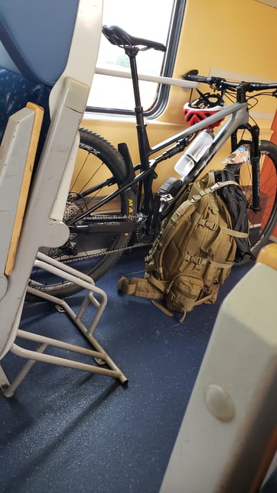 The image shows a bicycle parked inside a train car. The bicycle is a full-suspension mountain bike with a gray frame, black handlebars, and black wheels. A tan backpack is leaning against the bicycle, and a red and white helmet is attached to the handlebars. The train car appears to be empty, with blue seats visible in the foreground and a yellow wall in the background. The floor of the train car is a dark blue speckled material. The lighting suggests it is daytime, and the window in the background shows a blurred outdoor scene. The location is identified as Sučany, Slovakia.