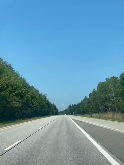 The image shows a highway stretching into the distance, flanked by dense green trees on both sides. The sky is a clear, bright blue, suggesting a sunny day. The road appears to be asphalt with white lane markings. The perspective is from a vehicle moving forward, as indicated by the vanishing point of the road. The overall scene evokes a sense of travel and open space, with the vibrant green of the trees contrasting with the gray of the road and the expanse of the blue sky. There are no people or specific objects visible that would indicate a particular event. The time of day appears to be daytime, likely late morning or afternoon due to the bright sunlight. The location is East Hawkesbury, Canada.