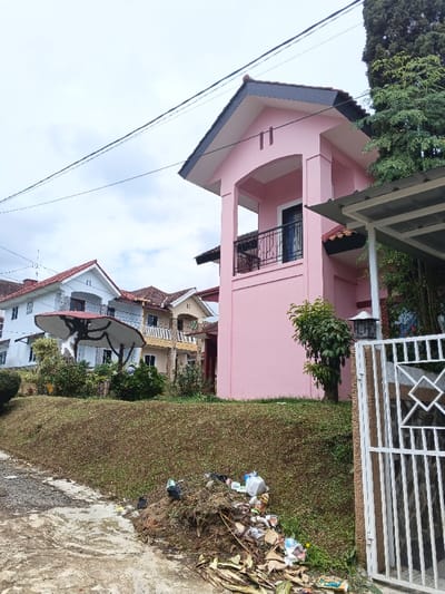 A pink two-story house with a balcony stands on a grassy hill. Other houses, one with a white exterior and a dark-colored shade covering an outdoor seating area, are visible nearby. A large pile of trash, including plastic bags and discarded items, sits at the bottom of the hill near the road. A white metal gate is partially visible in the foreground. Overcast sky is above.