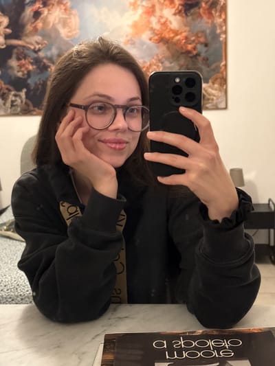 A young woman is taking a selfie with a black smartphone. She is wearing round glasses and a black sweatshirt. Her left hand is supporting her chin. Behind her is a painting, likely a reproduction of a classical scene with figures in a cloudy, celestial setting. To her right, a book with the title "WOOL" is visible. The setting appears to be indoors, possibly a residential space. There is no information provided that would indicate the city of Perugia, Italy as the location.