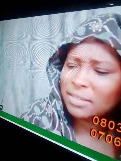 A woman wearing a patterned hooded garment is centered in the frame. Her eyes are closed, and she appears to be in a state of distress or sadness. The background behind her is a pale, patterned surface. In the lower right corner, orange numerals "0803" and "0706" are visible, suggesting they are part of a phone number. The overall image has a slightly tilted perspective, suggesting it was taken of a television or screen. A horizontal green stripe runs along the bottom of the image.