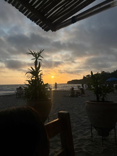 A view from under a wooden slatted roof on a beach in Montañita, Ecuador. The beach is sandy, and the sun is setting behind a group of people, silhouetted against the horizon. Several people are standing on the sand, and a group of three are seated further back. The sky is cloudy and overcast, but the sunset is visible in the distance. The scene is framed by two large potted plants. The location is consistent with coordinates -1.821 latitude and -80.756 longitude. The wooden slatted roof is likely part of a restaurant or bar, as a blue canopy with the word "Soda" printed on it can be seen in the background. A wooden chair is in the foreground of the image.