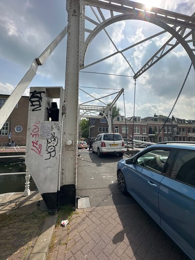 The image captures a scene on a bridge in Utrecht, Netherlands. The bridge appears to be a drawbridge, recognizable by its white, arching metal structure and chain mechanism. Cars are visible on the bridge, including a light blue car in the foreground and a white van with the license plate "38-HXF-8" further along. Buildings line the street in the background. Graffiti is present on the bridge's structure, including the word "ZOER" and other tags. A poster is attached to the bridge. The bridge also contains graffiti including a note that reads: "A FAILURE TO ENGAGE IN A WAR THAT ONLY CAN BE RESOLVED THROUGH INTERVENTION IMPLIES A HOPE FOR PEACE THROUGH NEGOTIATIONS"