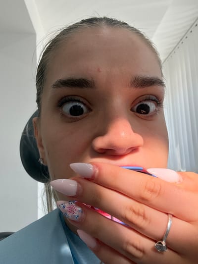 This media file is a close-up, rotated photograph of a person's face, focusing on their eye, nose, and hand with manicured nails. The individual appears to be young, with dark, well-defined eyebrows and large, expressive eyes that are looking directly at the camera. A strong shadow is cast across the left side of their face, suggesting a light source from the right, possibly indicating daytime.
The hand prominently displayed features long, stiletto-shaped fingernails painted in a pale pink or nude shade. One nail on the ring finger is decorated with a delicate floral design featuring blue petals and pink accents, with a small pink embellishment in the center. The person is wearing a simple silver ring with a clear stone on their ring finger.
The background is out of focus but appears to be an indoor setting with white, horizontal surfaces, possibly blinds or a textured wall, and a hint of a light blue garment or surface. A dark object, possibly a headrest or a hat, is visible behind the person's ear. The overall impression is intimate and personal, capturing a moment of self-presentation or a captured detail of their appearance. There is no visible text in the image.