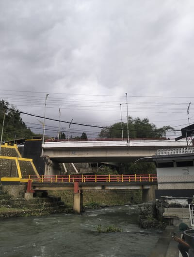 The image depicts a scene in the city of Blang Bebangka, Indonesia, featuring two bridges crossing a river. The foreground shows a murky, fast-flowing river with some aquatic vegetation. To the right of the river, a person wearing a green cap is seated, holding a phone and a cigarette. Adjacent to the riverbank is a structure with a tiled facade and a sign that reads "TEMPAT WUDHU" (ablution area).
A smaller, older bridge with red and yellow railings spans the river closer to the viewer. This bridge is supported by concrete pillars. Behind this bridge, a larger, more modern bridge with a white concrete surface is visible. This bridge also has red railings and is supported by substantial concrete piers. The sides of the embankment leading up to the larger bridge are reinforced with stone and yellow-painted concrete. Power lines crisscross the sky above the bridges. Trees and foliage are present on the hillsides in the background, and the sky is overcast with grey clouds.