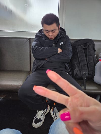 An image captures a moment inside what appears to be a moving vehicle, such as a train or bus, during an overcast day. The primary subject is a person sleeping soundly on a dark-colored bench seat, their head resting on their arm. They are dressed in a black hooded jacket with a white logo, possibly reading "EWE" or "3WE," and black pants. White socks and distinct sneakers with white laces and black and orange accents are visible on their feet.

To the right of the sleeping person, a hand extends into the frame, making a peace sign. This hand features bright, contrasting nail polish: fuchsia pink on the middle finger and vibrant orange on the ring finger. This hand belongs to another individual, whose blue-jeaned thigh is partially visible in the bottom right corner.

Behind the sleeping person's head, a black tactical-style backpack with multiple buckles and straps is placed on the seat. A grey headrest or small pillow is also partially visible. A large window runs along the side, revealing a blurry, light grey, and possibly rainy or foggy outdoor scene, suggesting dim daylight. The overall atmosphere conveys a sense of travel and rest.