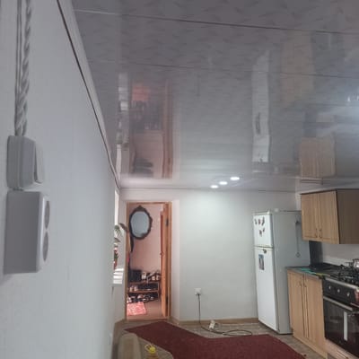 The image depicts an interior scene of a dwelling in Cepeleuți, Moldova. In the foreground left, a textured white wall is visible with two light-colored electrical outlets mounted vertically. A braided white electrical cord runs from the upper outlet and down towards the bottom of the frame. The ceiling above is a reflective, patterned white material.
The midground reveals a doorway leading into another room. This doorway is framed by light wood. Inside this room, an oval mirror with an ornate dark frame hangs on the wall. To the right of the doorway, a wooden shelf unit is filled with numerous pairs of shoes. A small, patterned rug lies on the floor in front of the shoe rack.
Further into the background and to the right, a kitchen area is visible. A white refrigerator stands against the wall. Adjacent to it are light-colored wooden kitchen cabinets and a stovetop with an oven beneath. The floor appears to be tiled or linoleum. Recessed circular lights are present in the ceiling. The overall impression is that of a modest, lived-in interior space. No people are visible in the image.