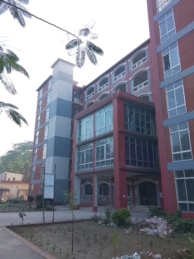 A multi-story building with a mix of red brick and grey-painted sections is shown. A modern glass-enclosed section protrudes from the center. Windows are numerous throughout the building. The foreground features a dirt patch with sparse vegetation. Tree branches are partially visible in the top left corner. The sky is overcast.