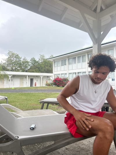 A medium shot captures a male seated on a grey lounge chair, appearing to be in the city of Sainte-Anne, France. The man has curly hair and is wearing a white tank top and red shorts. He is looking down, with his right hand resting on his right thigh.

The setting appears to be an outdoor patio or deck area. Behind the man, there is a building with multiple windows and some flowering plants. A grassy lawn and other trees are also visible in the background. A black cylindrical object sits on the chair next to the man. A white gazebo structure is partially visible on the left side of the frame. The overcast sky suggests it could be a cloudy day.