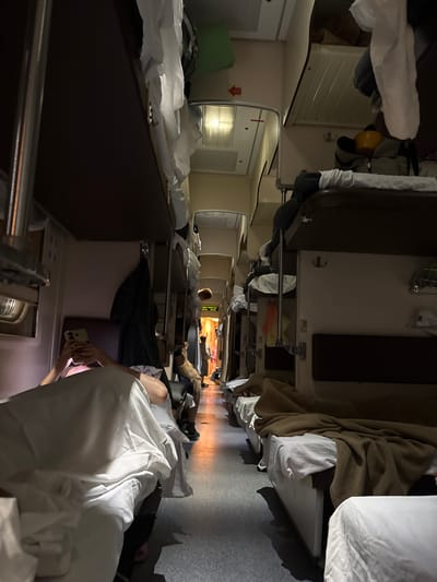 The image shows the interior of a compartment on a train, likely a sleeper car, with bunk beds lining the walls. In the foreground, a person is lying on a lower bunk, holding a smartphone. The compartment is dimly lit, with some overhead lights and light coming from further down the train car. Several beds are made up with white linens and brown blankets. Clothes are hanging on hooks and draped over bunks. The overall environment suggests a nighttime journey. The location is identified as Inza, Russia, which is a city in Penza Oblast, Russia, known for its railway junction.