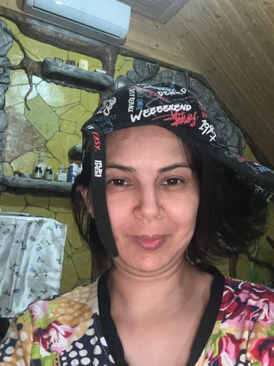 A woman is captured in a close-up selfie, looking directly at the camera with a slight smile. She is wearing a black bandana with white and red lettering that appears to say "WEEKEND" and "1997". Her attire includes a colorful, floral-patterned top with a black trim. The background suggests an indoor setting with a wall decorated with textured, stone-like elements and a yellow, stone-patterned wallpaper. A shelf is visible on the wall, holding several bottles. An air conditioning unit is mounted high on the wall. The scene is illuminated by artificial light. The image does not provide enough context to determine the specific location within Kalininskaya, Russia.