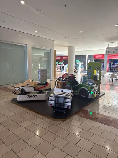 This media file captures a section of an indoor shopping mall in National City, United States of America. The primary subjects are a row of coin-operated children's rides, shaped like miniature vehicles. These include what appears to be a green jeep with "ALIEN PATROL" written on it, a red car, and a white and black striped vehicle with a plush dog toy in the driver's seat. Another ride, a beige and red boat-like vehicle, is prominently displayed with "TOM CAT" in bold lettering on its side.
The rides are positioned on a dark, raised platform. In the background, there are glimpses of stores and mall interiors, including a prize redemption area with colorful balls and a sign that says "QUICKY PRIZES." A directional sign pointing to various stores like "JCPenney," "Macy's," "AMC," and "ROUND1" is also visible.
The scene appears to be set during the daytime, with artificial lighting illuminating the indoor space. The floor is tiled with a light-colored stone pattern and features a decorative strip of reddish-brown marble. The overall mood is casual and family-oriented, typical of a shopping mall environment. No specific activity or event is explicitly depicted beyond the presence of the rides, suggesting a normal operating day at the mall. The image is taken from a slightly tilted perspective, giving it a dynamic feel.