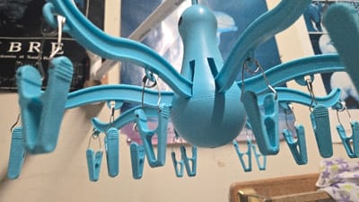 The image depicts an indoor residential environment, likely within a dwelling in Lompoc, United States, suggested by a partially visible poster referencing "University of California at Santa Barbara" on the left wall. The primary subject is a light blue, plastic, octopus-shaped clothes drying rack suspended from an unseen overhead fixture, positioned in the foreground and center of the frame. This rack features multiple radial arms, each terminating in a small metal hook or ring from which individual light blue plastic clothespins are attached. No garments are present on the clothespins.

In the background, a light-colored wall is visible. To the left, a dark poster with white text is partially obscured by the drying rack; legible fragments include "B R E" and "ifornia at Santa B". Further to the right in the background, another lighter-toned image or poster with abstract blue shapes is visible. In the lower right quadrant, a section of wooden furniture, possibly a dresser or headboard, is present, topped by a folded piece of fabric featuring a purple and white floral pattern. No individuals are depicted, and no active actions or direct interactions are occurring. The lighting suggests an interior setting with ambient illumination.
