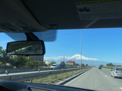 The image shows a highway scene viewed from inside a car. The road is straight, with multiple lanes in each direction, and a silver car is visible on the right. Other cars are driving further down the road. A metal guardrail lines the side of the road, with grass and some buildings beyond. In the distance, a large snow-covered mountain is visible against a clear blue sky. The car's rearview mirror reflects a building and some greenery. The location is Ayntap, Armenia.