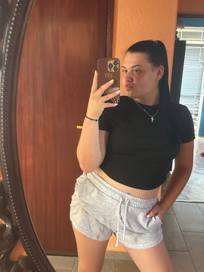 A young woman with dark hair in a ponytail, wearing a black crop top and gray sweat shorts, takes a mirror selfie indoors. She is holding a smartphone with a leopard print case. Her left hand is in her pocket, and her right arm is extended to hold the phone. A silver necklace with a cross pendant is visible around her neck. The background shows a wooden door on the left and an orange wall. The floor appears to be tiled. The image does not provide any specific clues about its location within the city of Rogosh, Bulgaria.