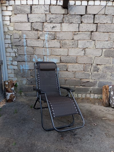 A dark-colored reclining chair stands on a paved surface in Кодинка, Russia. The chair is positioned in front of a light grey brick wall with some minor blue graffiti markings. To the left and right of the chair, at the base of the wall, are sections of cut wood. A partial white frame is visible on the left side of the image.