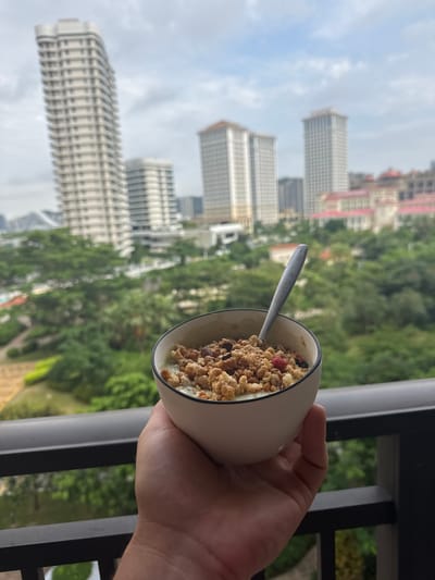 A hand holds a bowl of granola and yogurt on a balcony overlooking a city. The bowl is white with a black rim and contains granola with dried berries and possibly nuts. A silver spoon is resting in the bowl. In the background, several tall buildings are visible, including a prominent, curved white building. Lush green trees and other buildings can be seen below the skyscrapers. The sky is overcast. The image appears to be taken from a high vantage point, possibly a hotel room or apartment, offering a panoramic view of the urban landscape and surrounding greenery. The location is identified as Xiangzhou, China.