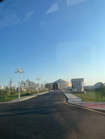 The image depicts a road leading towards a large, dome-shaped building with intricate patterns on its exterior, resembling a yurt. To the right of the road, there is a small, modern-looking booth with glass windows. The area is landscaped with neatly trimmed grass and young trees, some of which are bare. Ornate streetlights with multiple lamps line the walkway. The sky is clear and blue, with a few faint specks of pink and black that could be dust or distant birds. In the background, several other yurt-like structures are visible. The overall scene appears to be a modern, well-maintained public space or park in a city, likely designed with cultural or architectural influences from nomadic traditions. The location is identified as Қазақстанның 30 жылдығы, Kazakhstan.