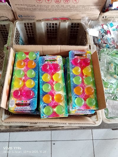 The image shows three packages of brightly colored jelly candies, each containing 12 pieces of jelly. The candies are arranged in four rows of three, with each row containing a different color. The packages are sitting on a brown cardboard box, which is on top of a white metal wire basket. The basket is partially filled with other packages of candies. The image is taken from a slightly elevated angle. The location is in Kecamatan Anjatan, Indonesia, at coordinates -6.416 latitude and 107.958 longitude. The environment is a small shop or market. The objects are three packages of jelly candies. The subjects are the candies. The actions are the candies being packaged and sold. The interactions are the candies being purchased by customers.