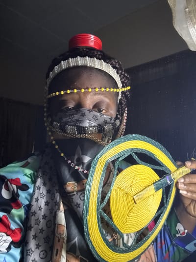 A person, seemingly female, is holding a round, woven fan. She is wearing beaded necklaces and a pearl headband, as well as a patterned face mask. She is surrounded by a collection of patterned fabrics, including those with cartoon characters and designer logos. The image's location is not discernible from the visual data provided. No location of Madangala, Nigeria is evident.