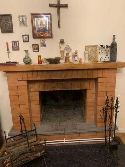 The image shows a domestic interior with a fireplace and its mantelpiece, located in Mepiskalaki, Georgia. The fireplace is constructed of orange bricks with a stone hearth. On the mantelpiece, there are several religious icons, a wooden cross mounted on the wall above, a vase, an oil lamp, and various decorative items including a candle holder with black metal accents, and a tall, dark-colored bottle. To the left of the fireplace, a metal log holder is filled with firewood. To the right, a set of black metal fireplace tools, including a poker, brush, and tongs, stands upright. The room appears to be dimly lit, with the primary light source possibly coming from a lamp or overhead fixture not visible in the frame.