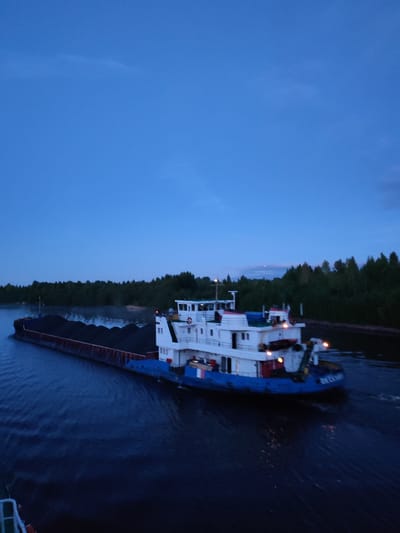 The image depicts a tugboat pushing a barge laden with a dark cargo, possibly coal, on a body of water. The tugboat is white with blue accents and has multiple decks. The barge stretches behind the tugboat, disappearing into the distance. Lush green trees line the shore on either side of the water, indicating a natural or semi-natural environment. The sky is a deep blue, suggesting twilight or dusk. There are no visible people in the image. The location is identified as Annensky Most, Russia.